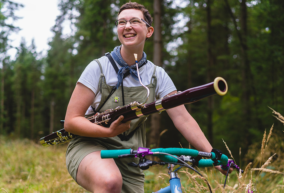 A photograph of participant of RCM Accelerate programme holds their bassoon while sitting on a green bike, with grass and trees visible in the background.